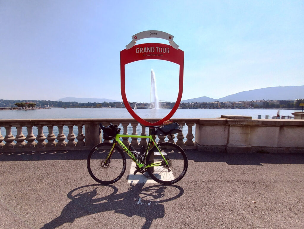 Grand Tour of Switzerland sign with a bicycle and the Jet d'Eau fountain on Lake Geneva in Geneva, Switzerland