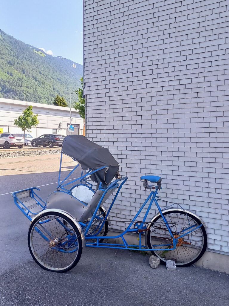 An old blue three-wheeled cycle rickshaw with a black canopy parked beside a wall in Interlaken.