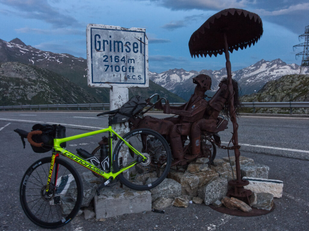 Road bike beside the Grimsel Pass sign and metal sculpture at 2,164 metres.