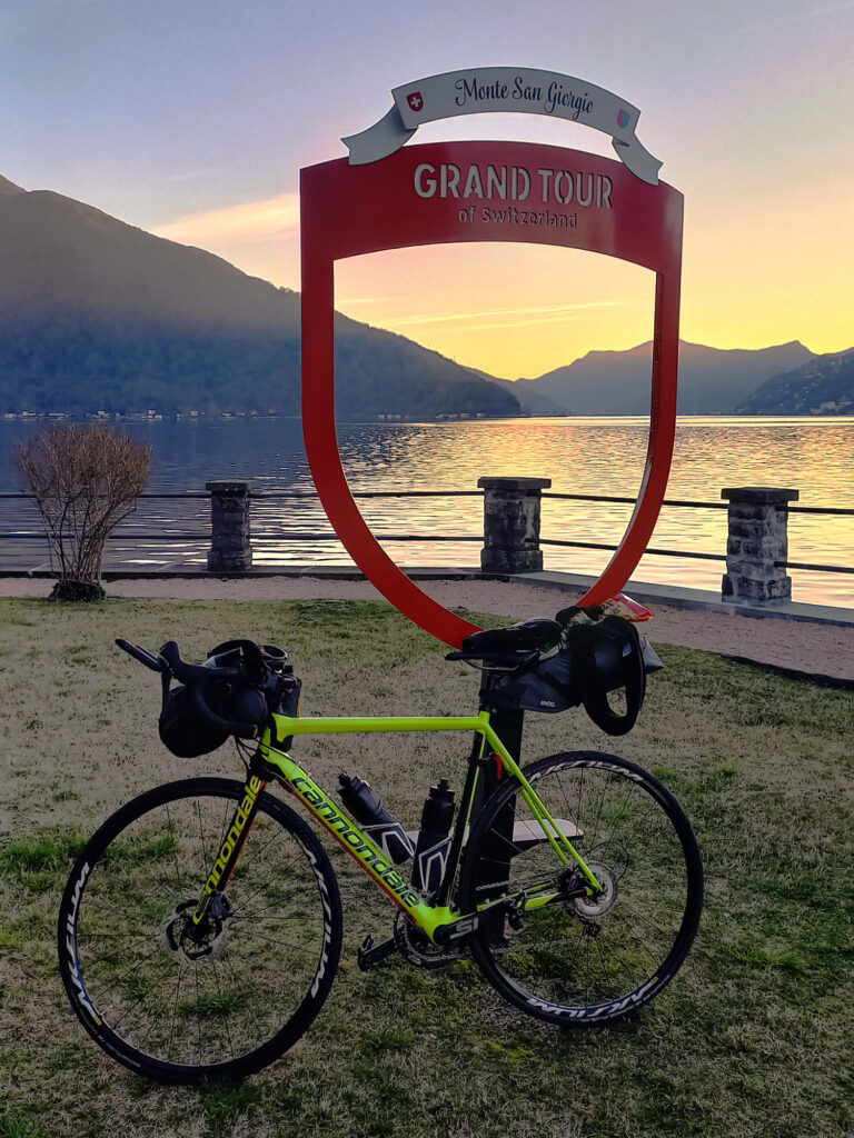 Cestný bicykel s batožinou pred znakom Grand Tour of Switzerland pri Monte San Giorgio