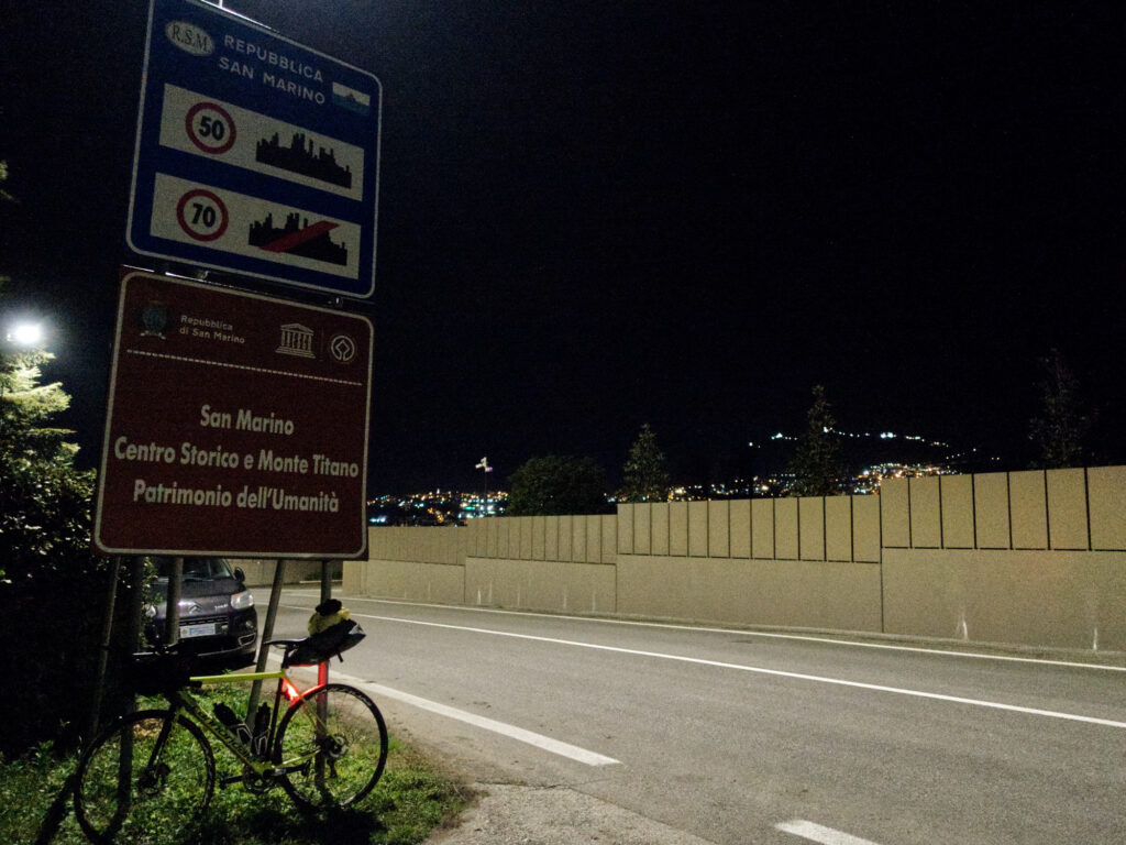 Bicykel v noci opretý o dopravné značenie na hranici San Marína s osvetleným mestom v pozadí