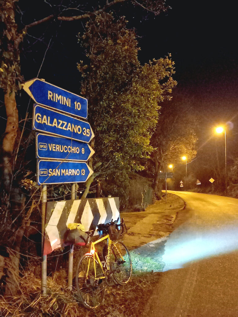 Bicykel opretý v noci pri smerovníku ukazujúcom posledných 13 kilometrov do San Maríno